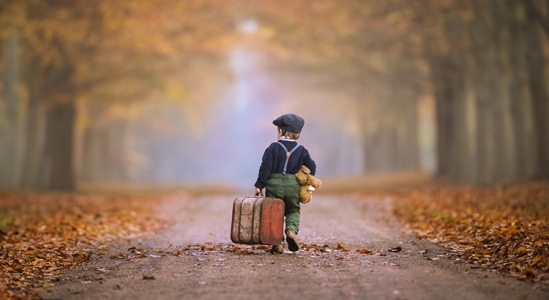 Small child holding a suitcase and teddy bear walking on a street dusted with leaves